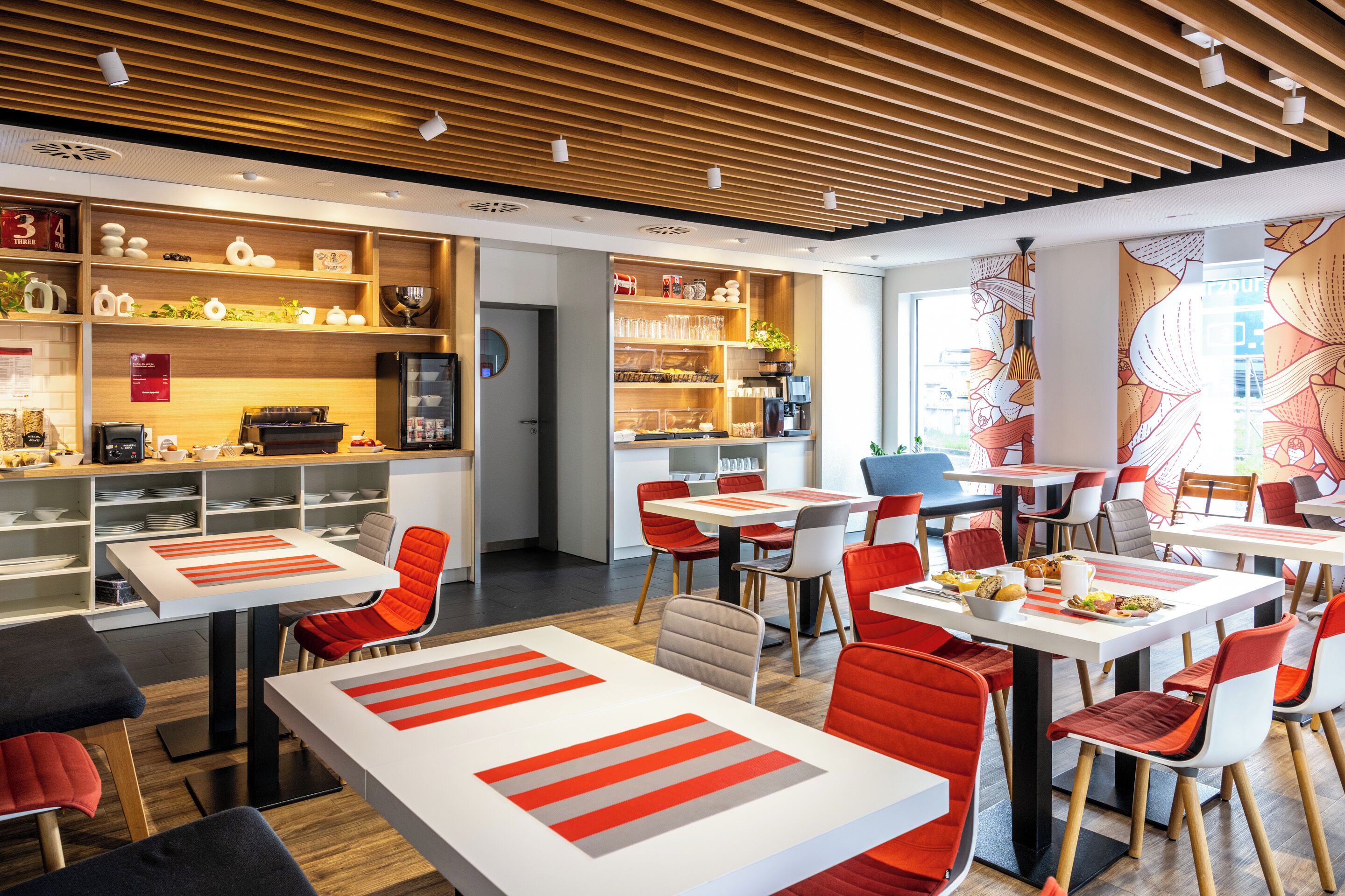 A modern restaurant with wooden ceiling slats, white tables with red-striped placemats, and red and white chairs.