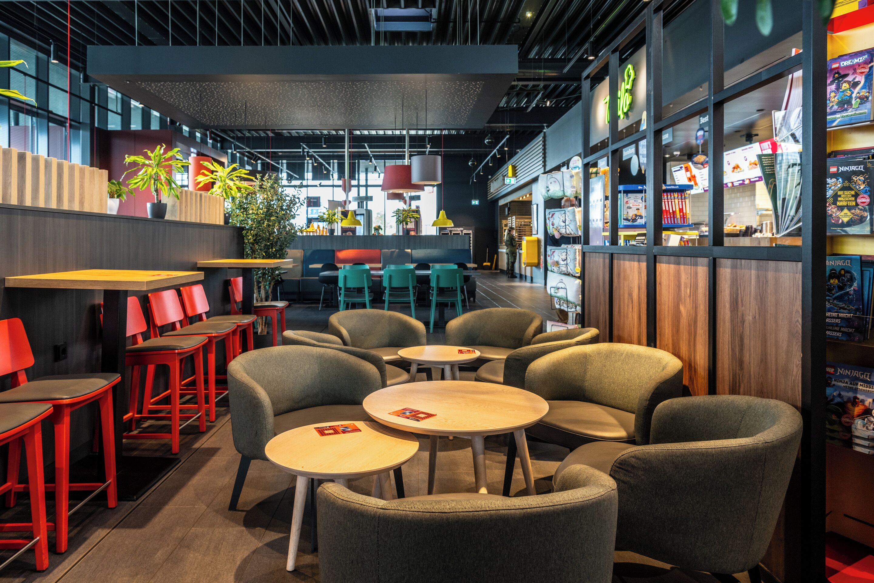 Interior de restaurante moderno con varias zonas de estar, incluyendo mesas redondas y sillas rojas, además de plantas y objetos decorativos.