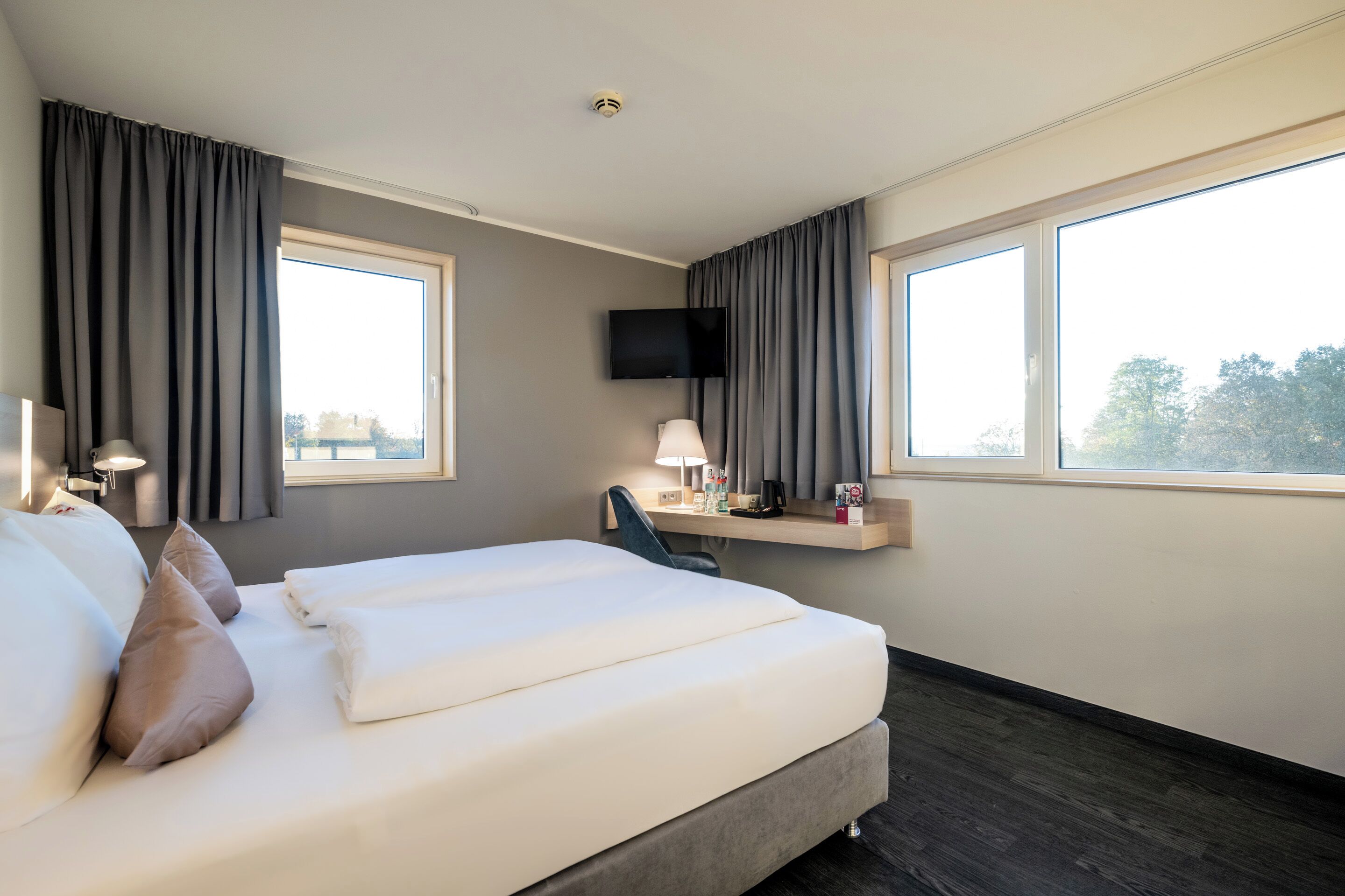 A hotel room with a bed, a desk, a chair, a TV, and large windows.