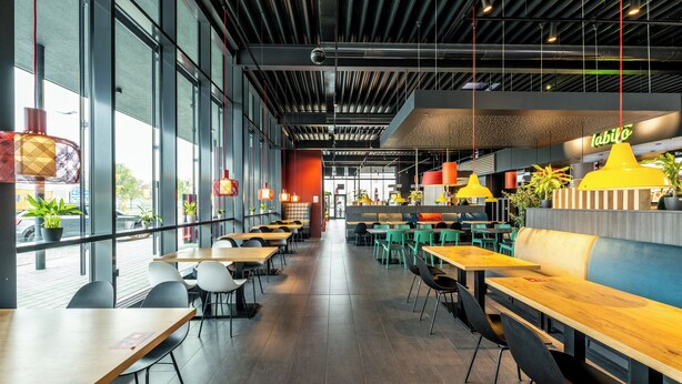 Interior moderno de restaurante con techos altos, vigas a la vista y ventanas amplias. Mesas y sillas dispuestas para comer.