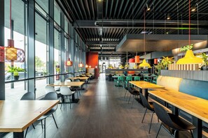 Un intérieur de restaurant moderne, caractérisé par de hauts plafonds, des poutres apparentes et de grandes fenêtres. Les tables et les chaises sont disposées pour accueillir les clients.