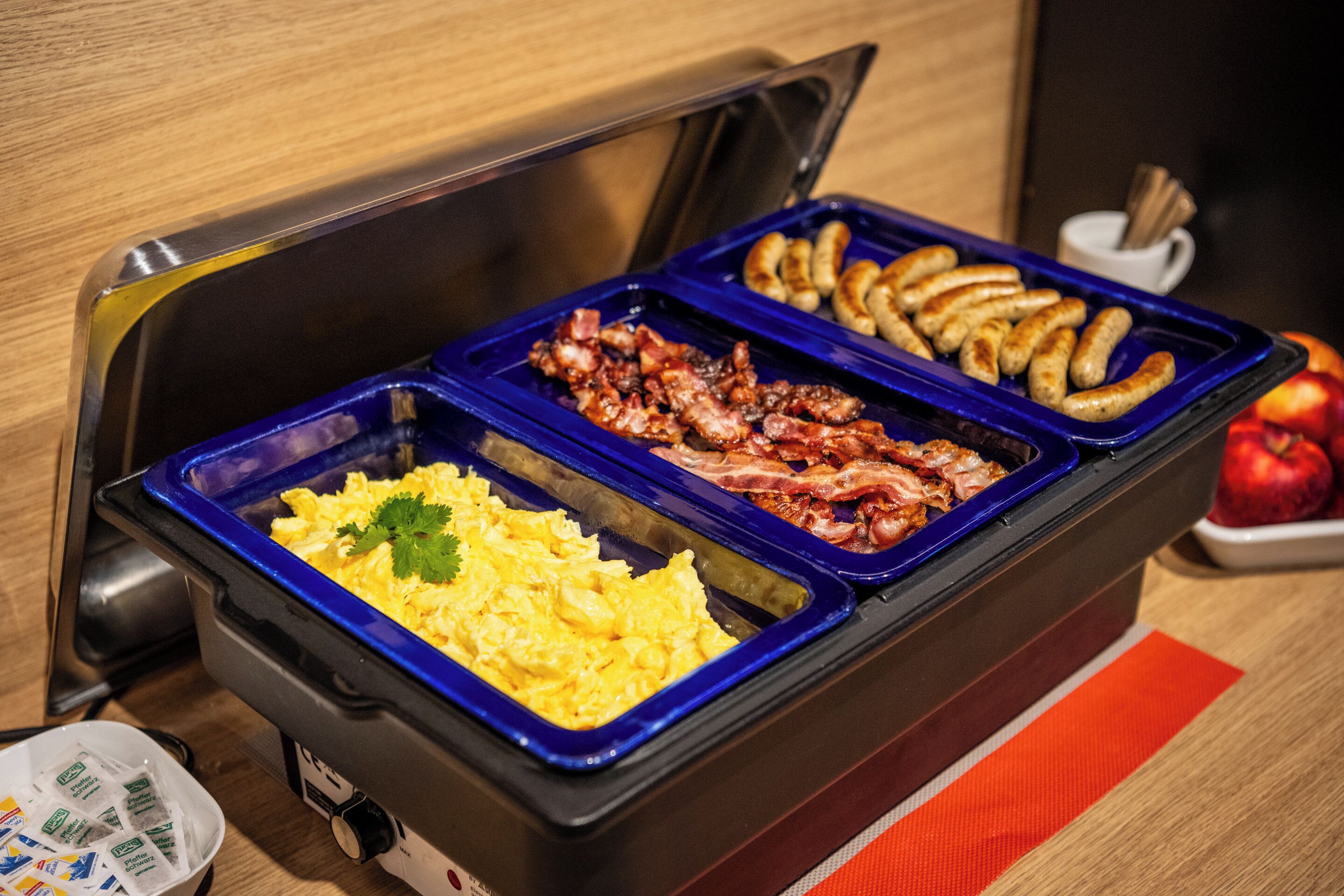 A buffet with scrambled eggs, bacon, sausages, and a fruit bowl.