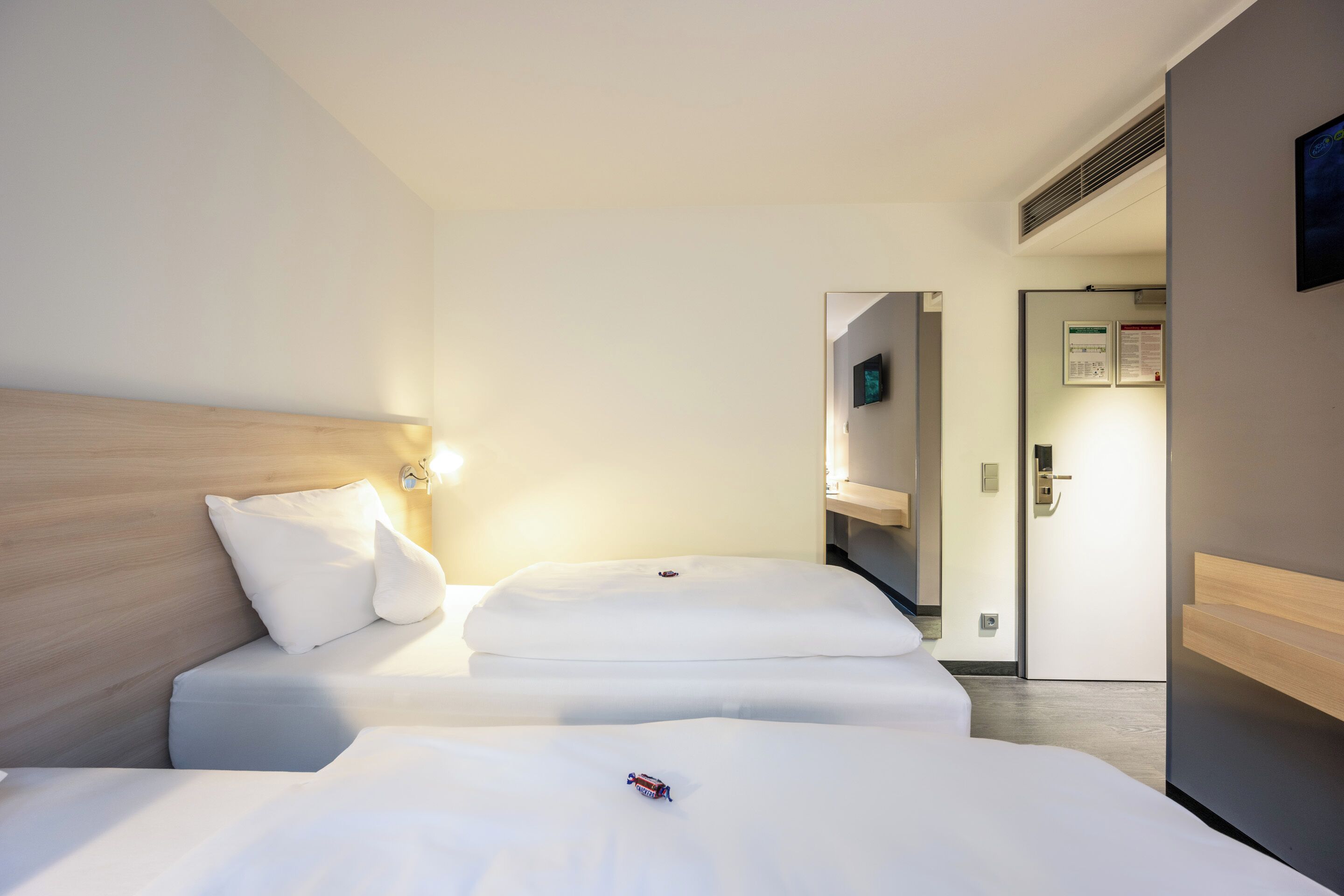 A hotel room with two beds, a wooden headboard, a wall-mounted TV, and a door leading to a bathroom.