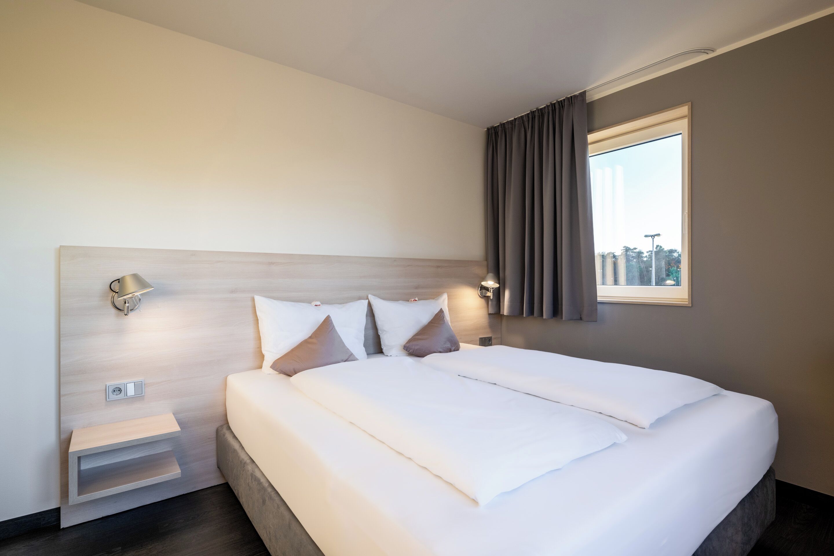 A modern hotel room with a large bed, a nightstand, a wall-mounted lamp, and a window with curtains.