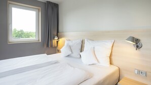 Una habitación de hotel con cama, ventana, lámpara de pared y mesita de noche.