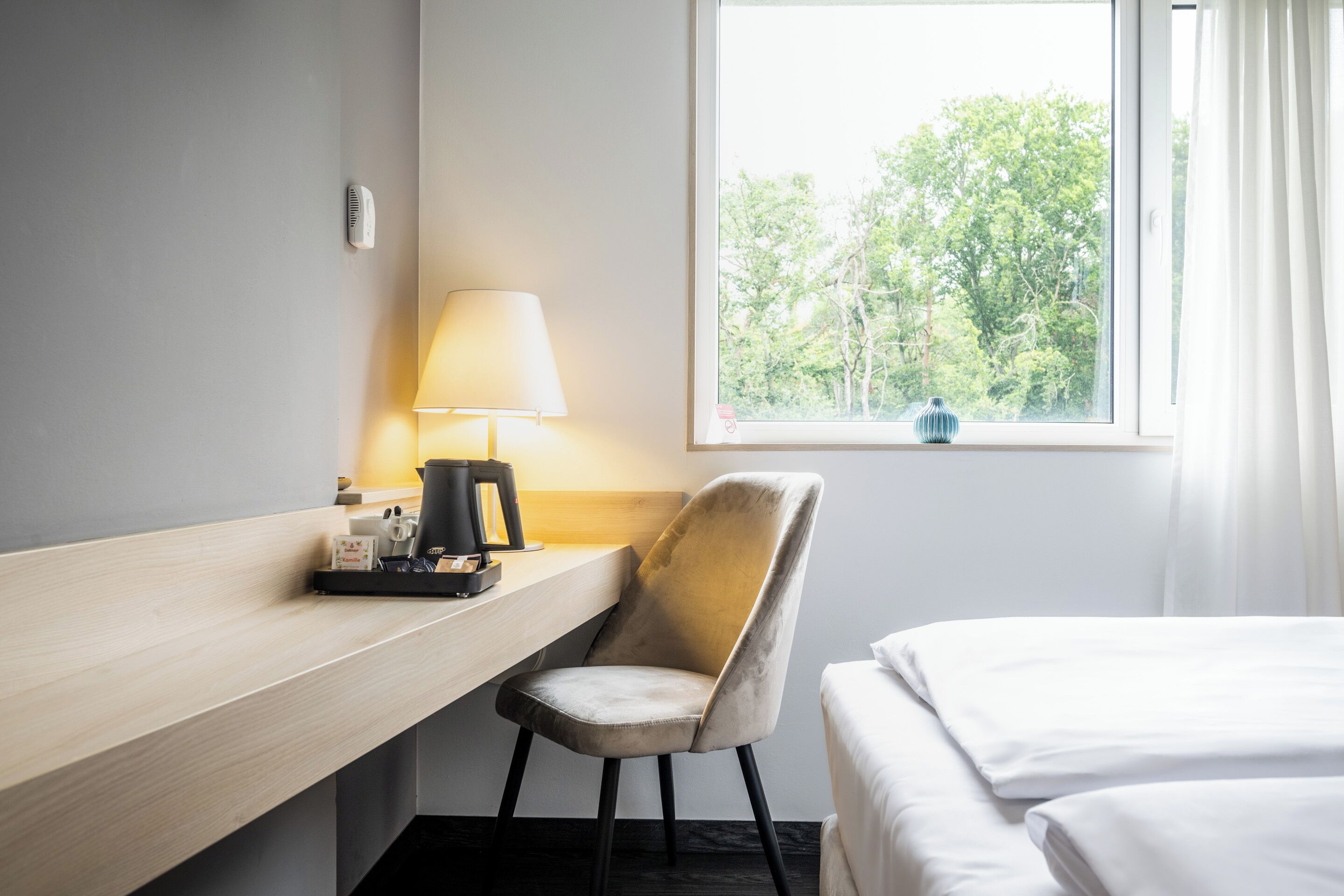 A hotel room with a bed, a desk with a lamp, a chair, and a window with a view of trees.