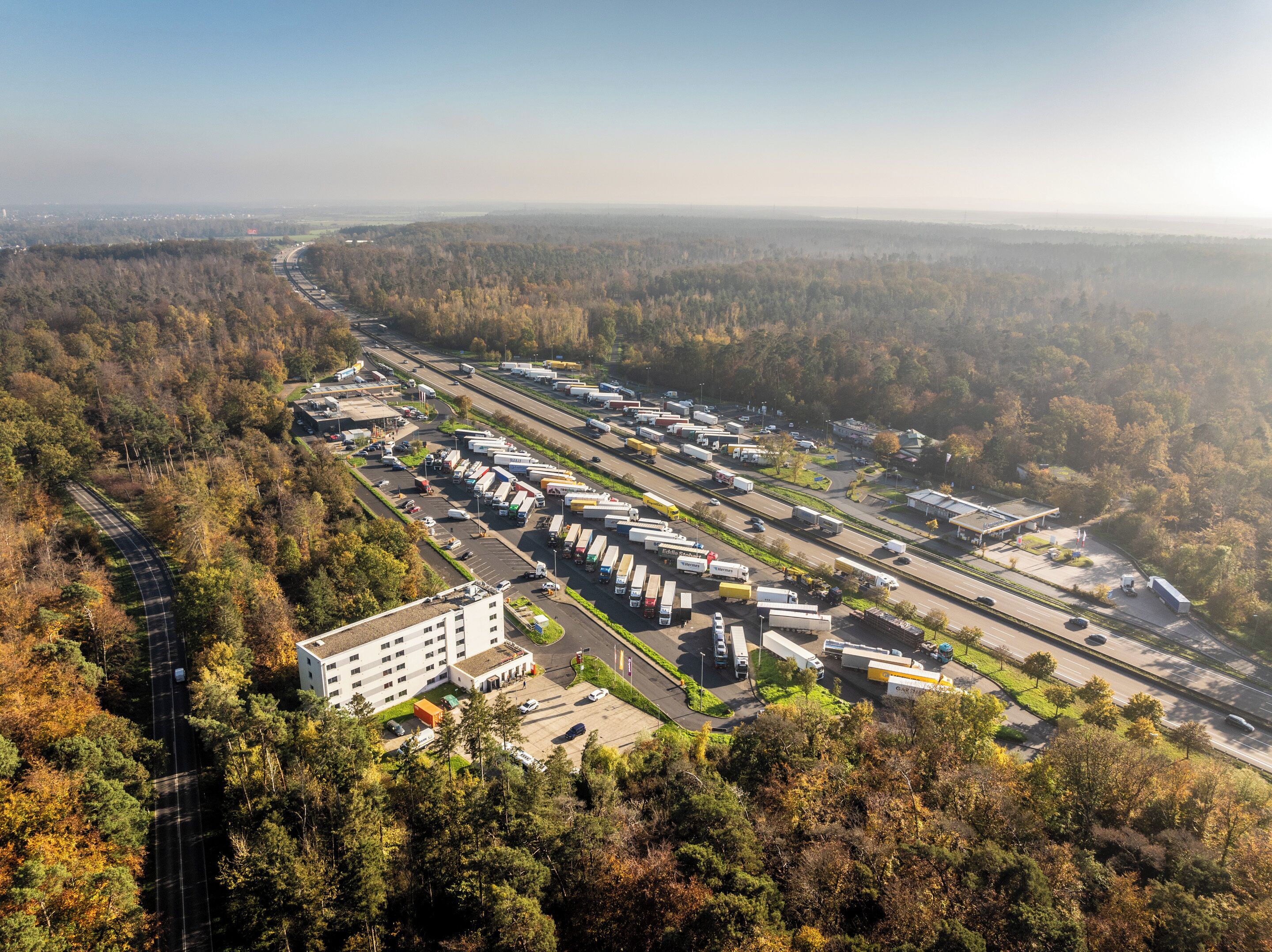 An aerial view of a logistics hub with multiple truck parking areas, a central building, and a highway in the background.