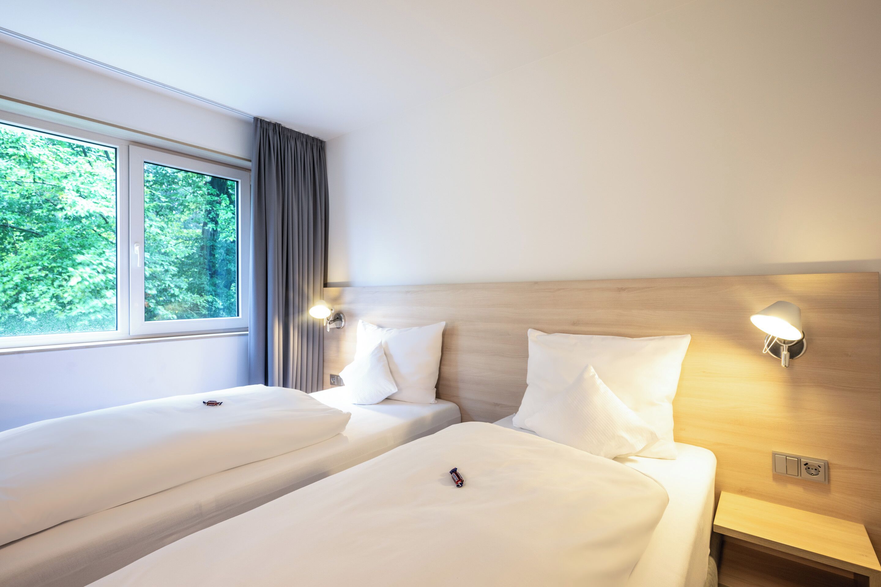 A hotel room with two beds, a window with curtains, a wall-mounted light, and a wooden headboard.