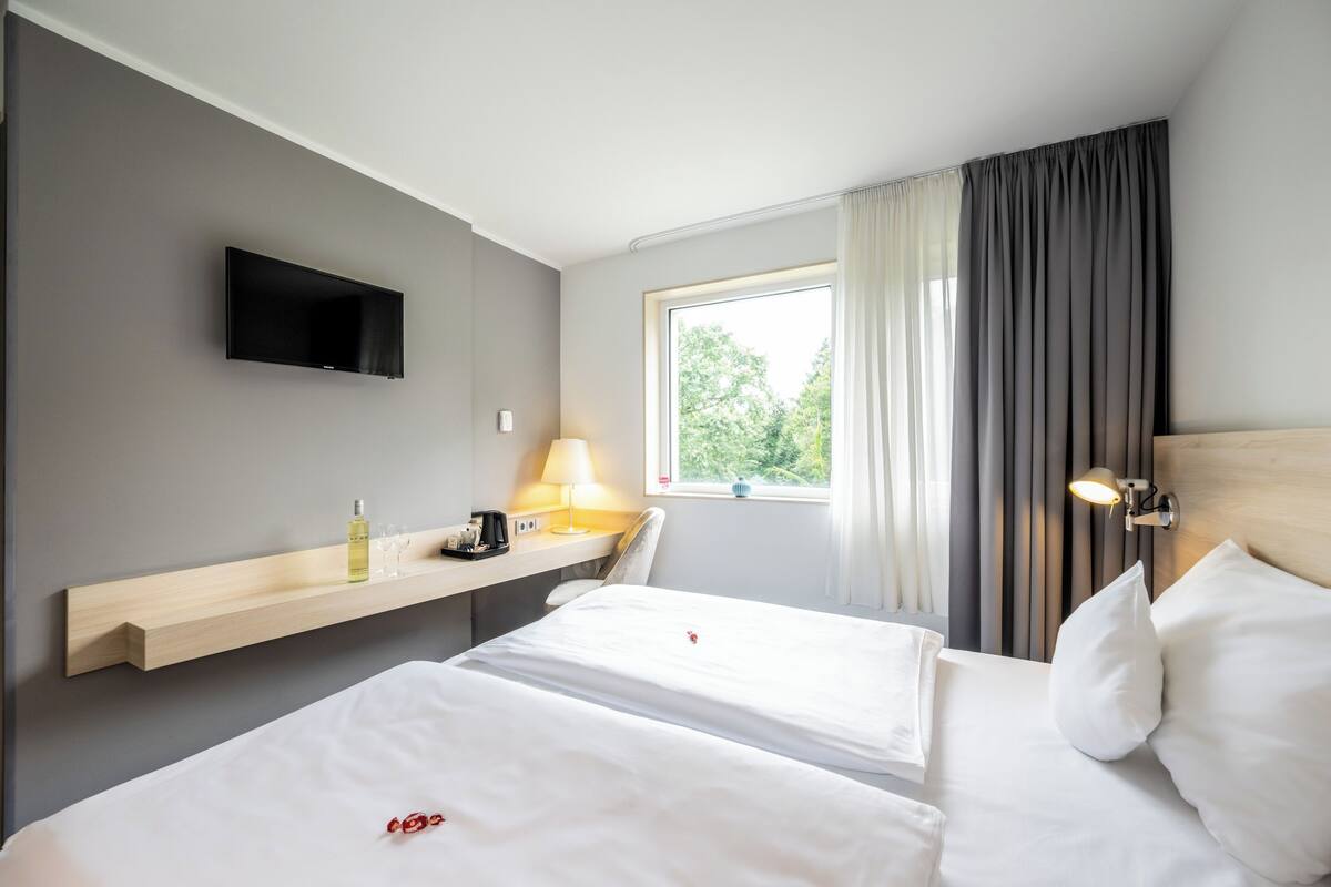 Hotellihuone, jossa on kaksi sänkyä, seinälle asennettu televisio, yöpöytä lampun kanssa ja ikkuna, jossa on verhot.