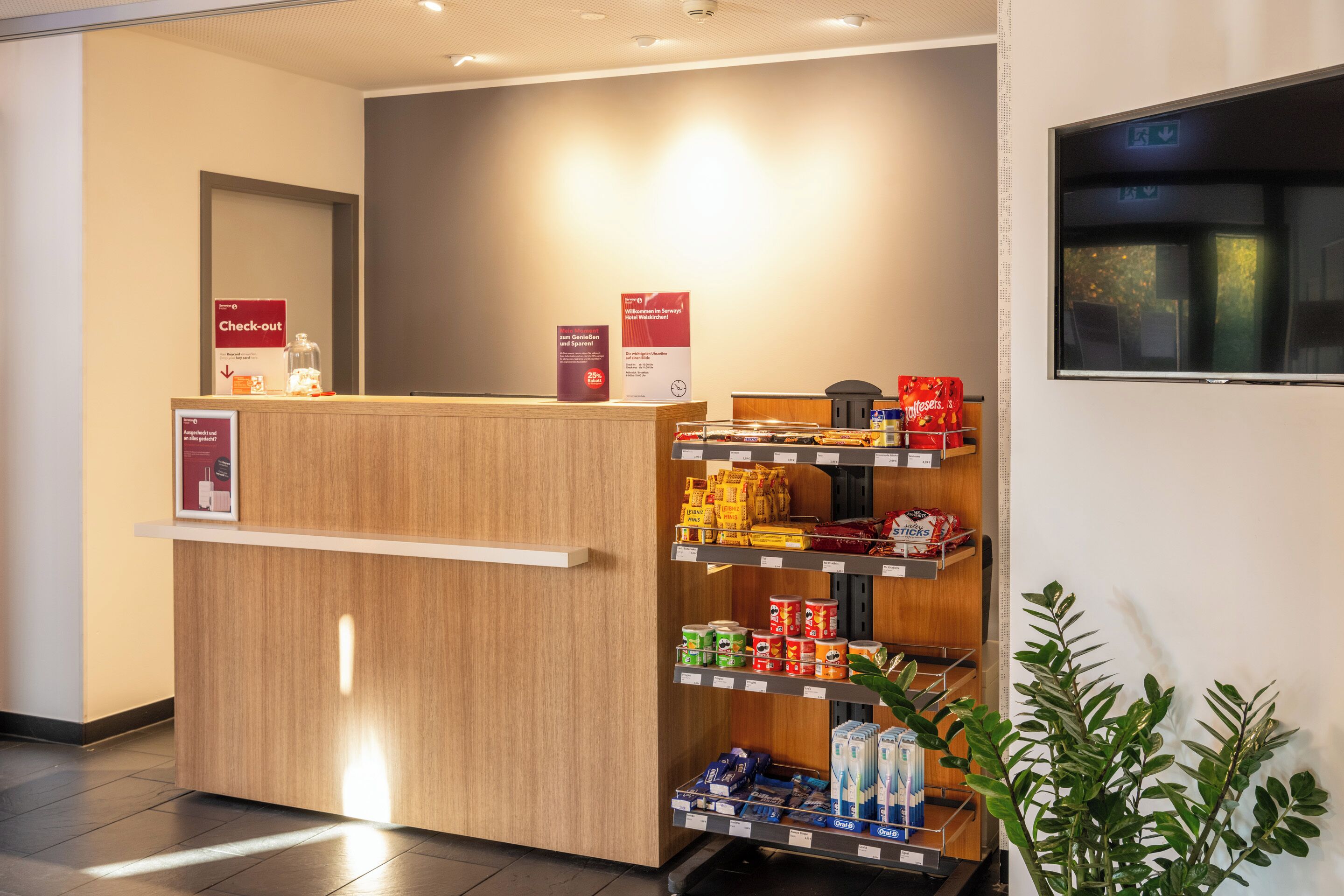 A reception desk with a sign indicating a check-out area, a shelf with various snacks, and a plant to the right.