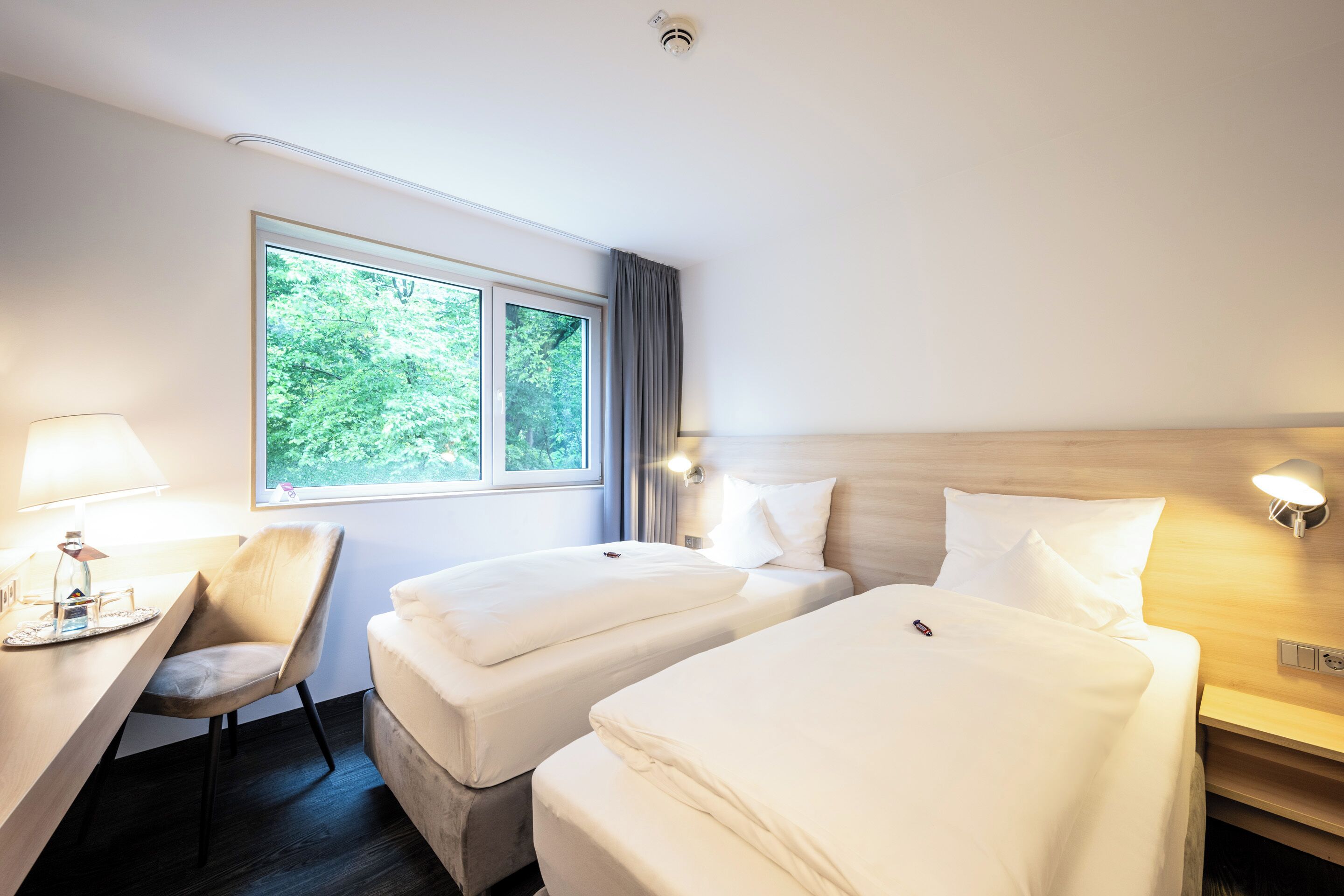A hotel room with two beds, a desk with a lamp, a chair, and a window with curtains.