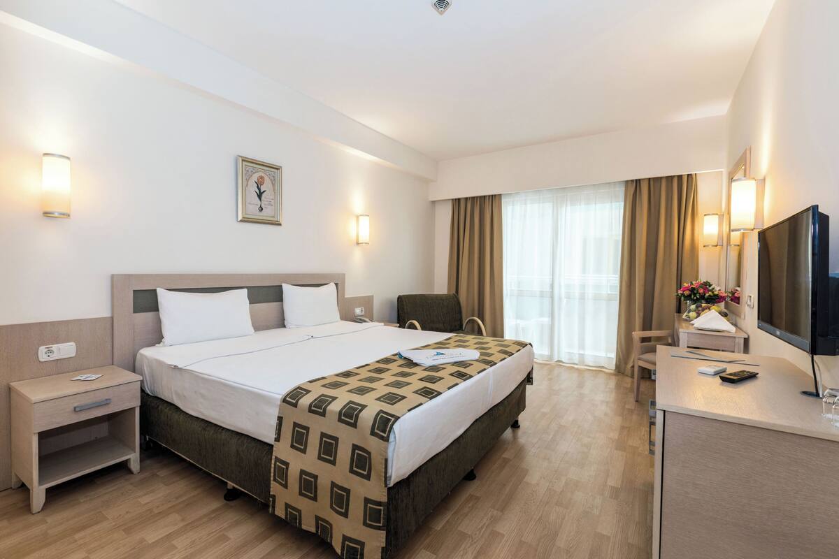 A hotel room with a large bed, bedside tables, a desk with a chair, a TV, and a window with curtains.