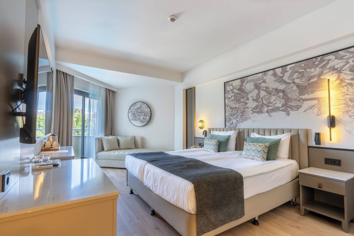 A modern hotel room with a bed, bedside tables, a desk, a television, and a large window with curtains.
