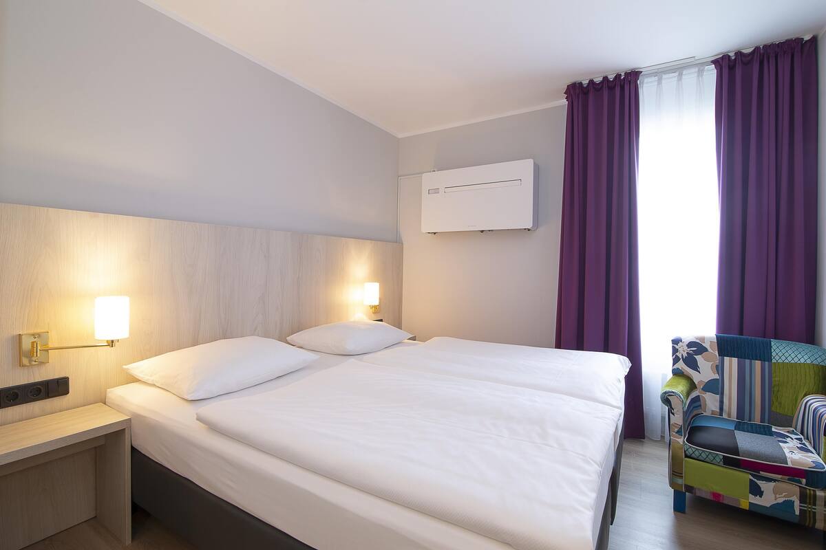A hotel room with a bed, bedside lamps, a window with curtains, and a chair.