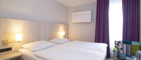 Ein Hotelzimmer mit Bett, Nachttischlampen, einem Fenster mit Vorhängen und einem Sessel.