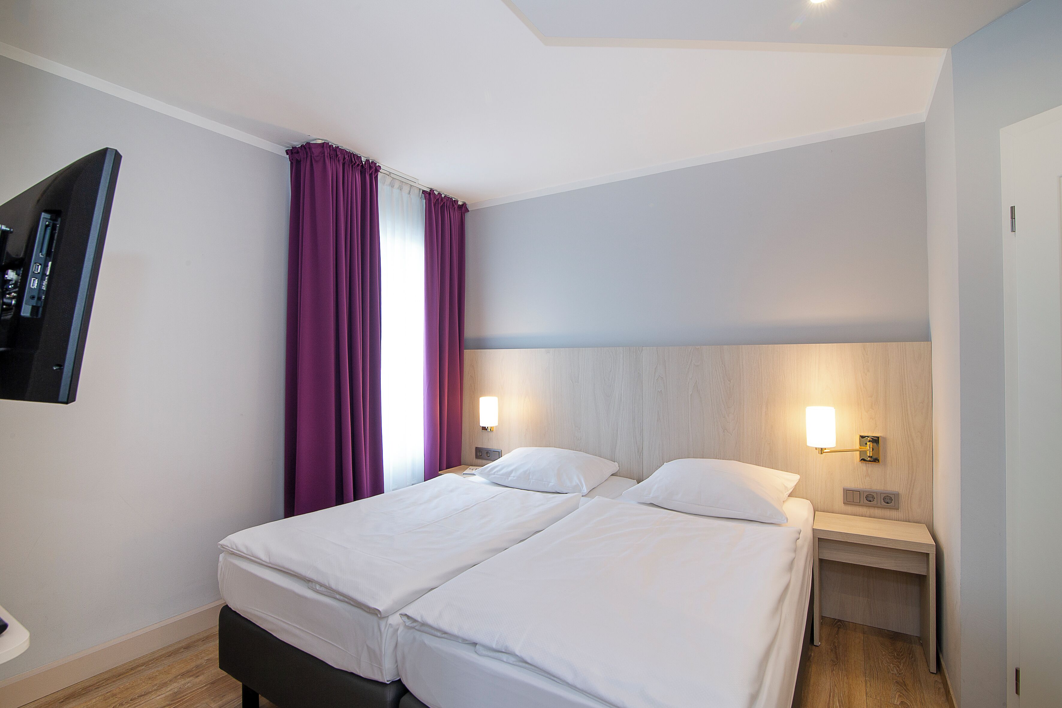 A hotel room with two beds, a flat-screen TV mounted on the wall, and a nightstand with a lamp.