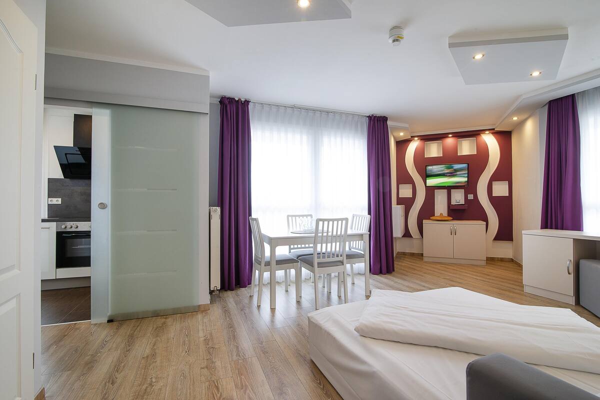 A hotel room with a bed, a desk with a computer, a TV, and a dining area with chairs and a table.