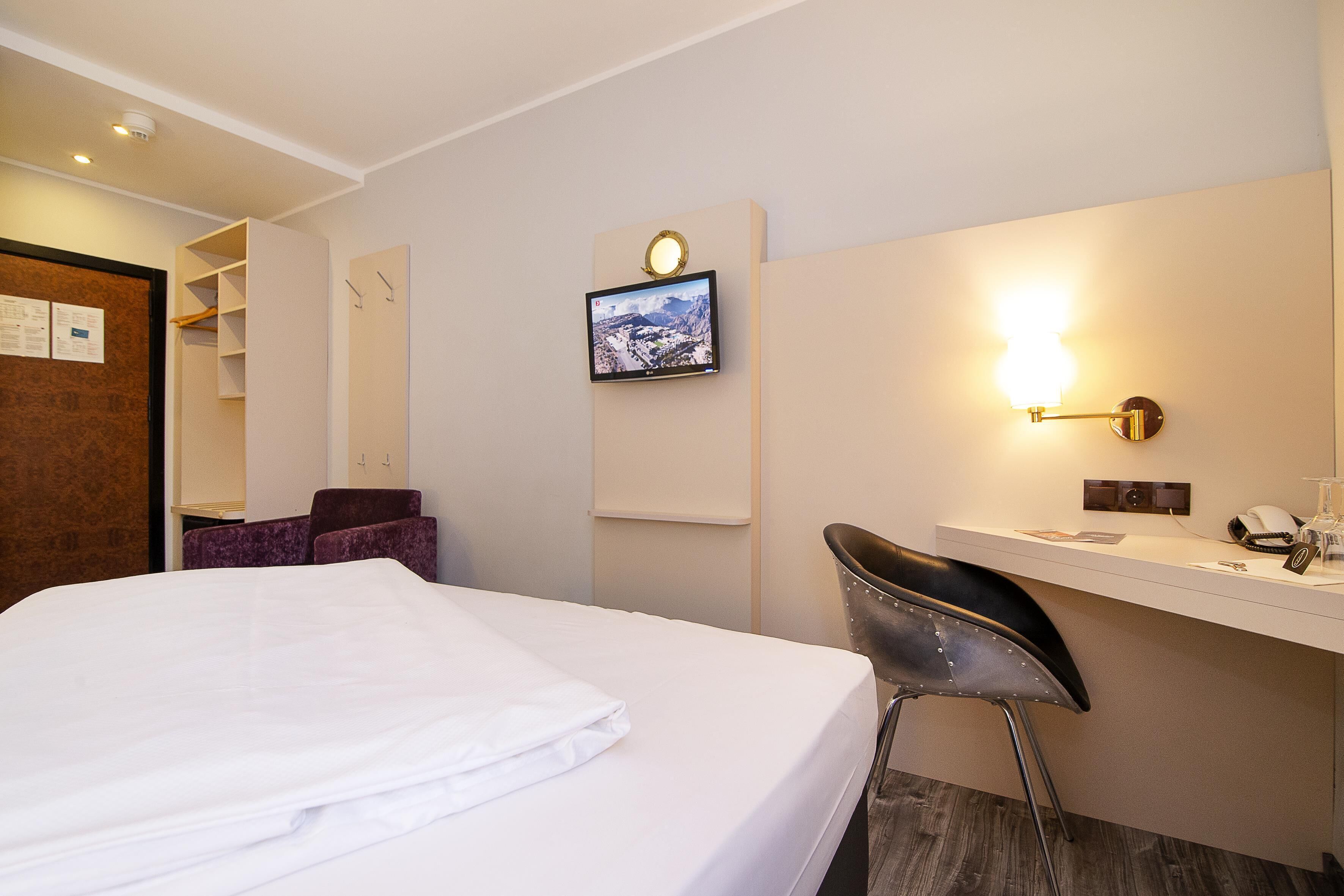 Ein Hotelzimmer mit Bett, Schreibtisch mit Stuhl, einem angebrachten Fernseher und einer Wandleuchte.