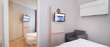 A modern hotel room with a bed, a gray armchair, a flat-screen TV mounted on the wall, and a built-in air conditioning unit.