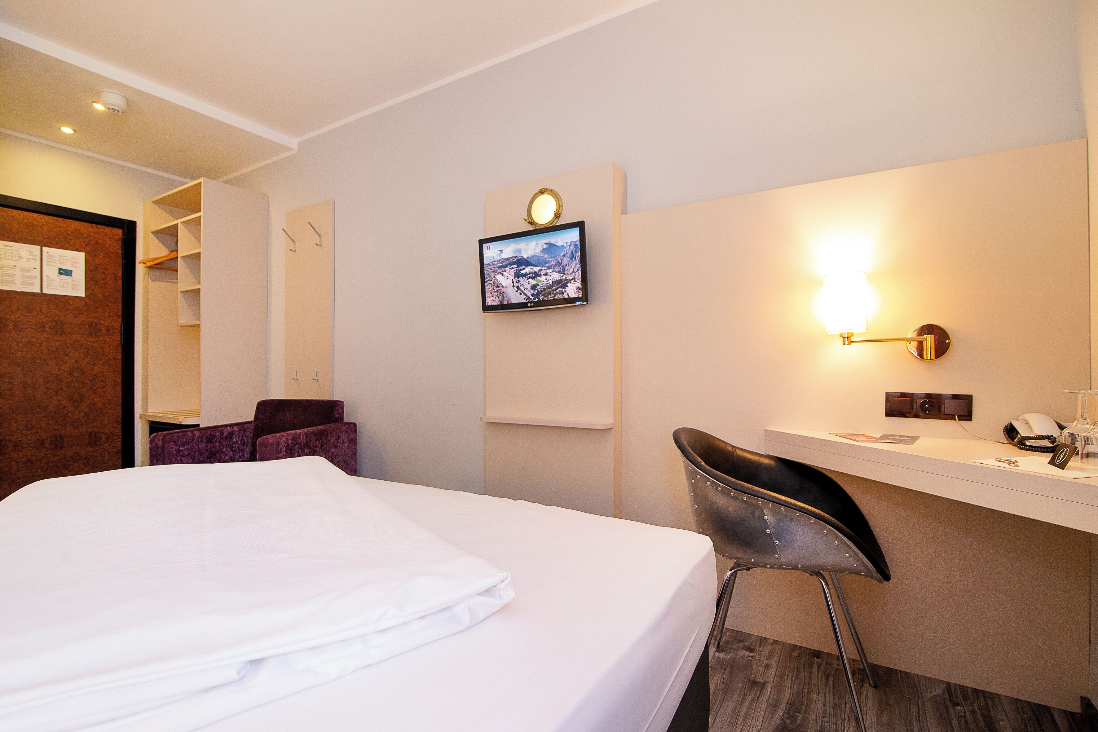 A hotel room with a bed, a desk with a chair, a TV mounted on the wall, and a wall-mounted light fixture.