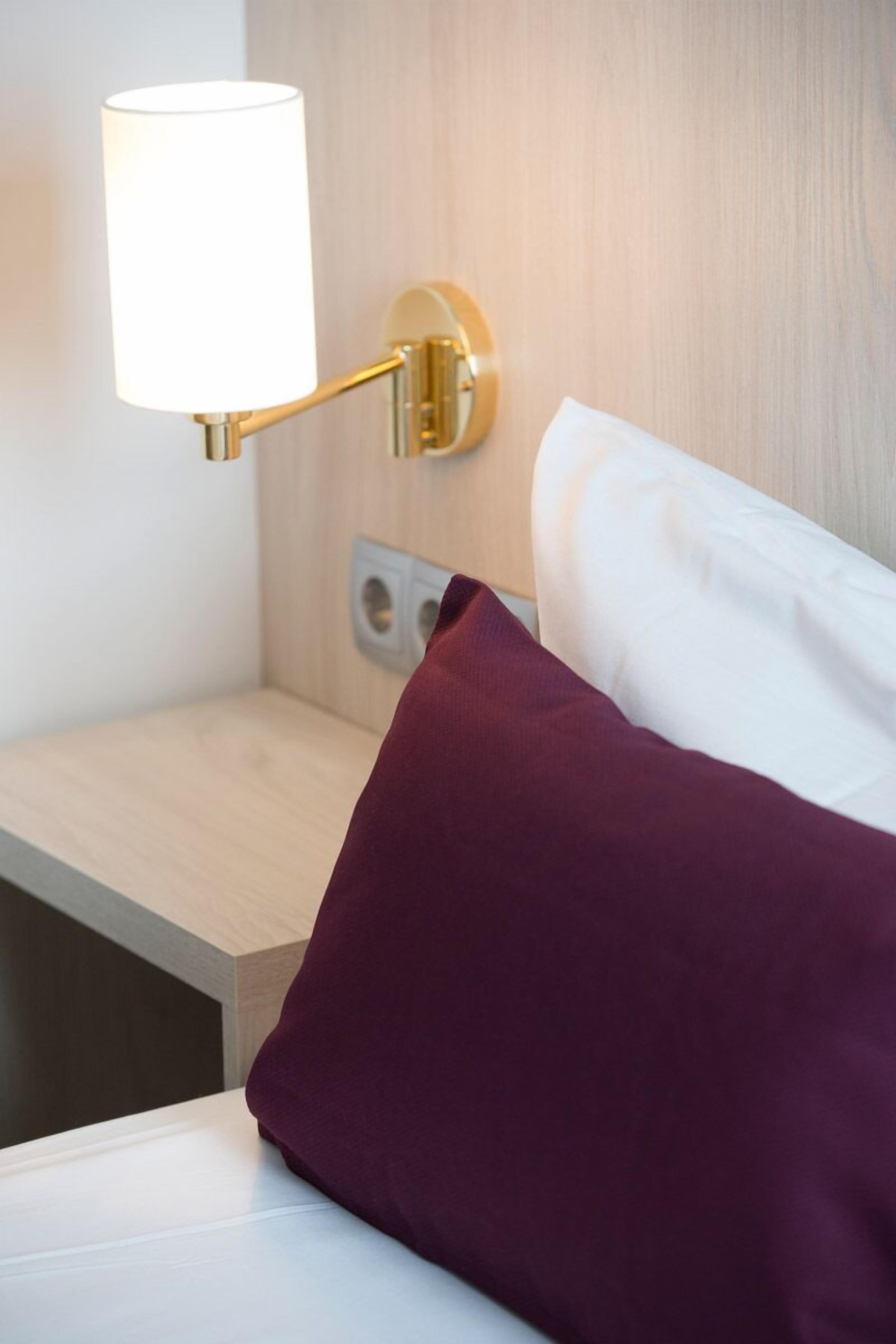 Una cama bien hecha con un almohadón blanco y uno burdeos, una mesita de noche con una lámpara, e interruptor en la pared.