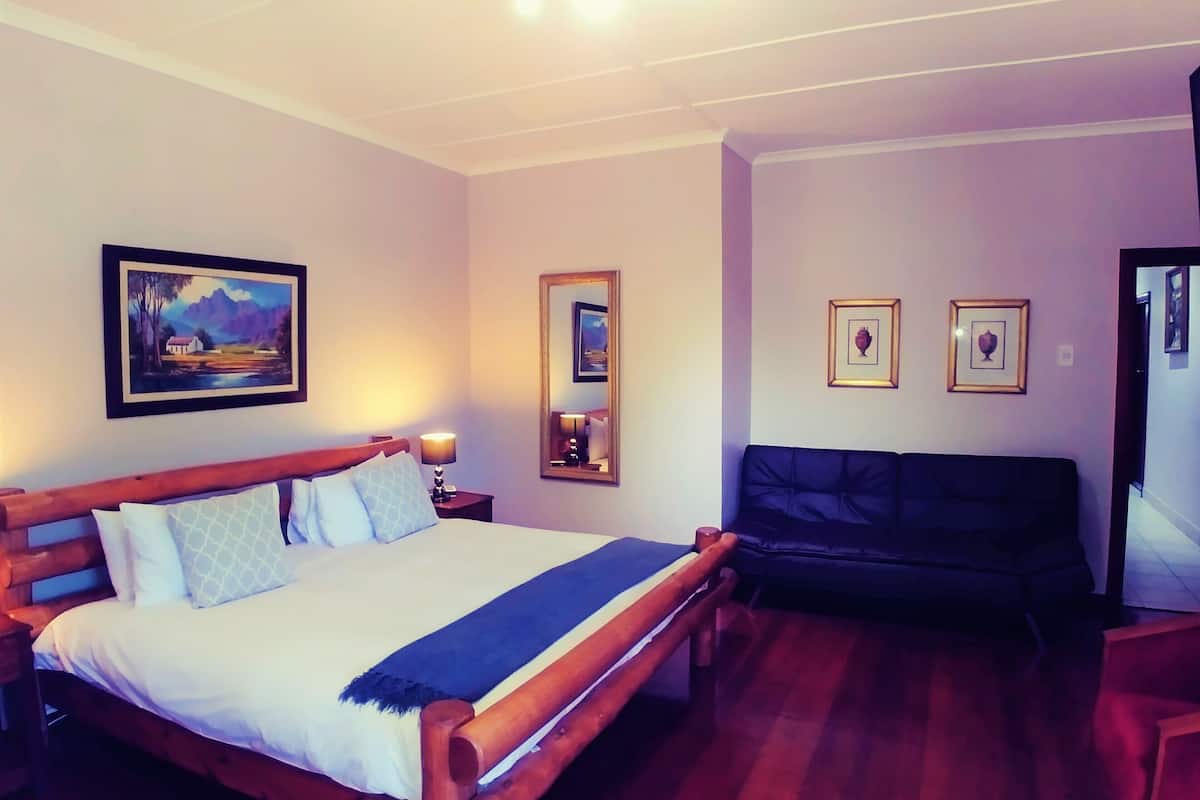 A hotel room with a wooden bed, a sofa, a nightstand with a lamp, and two framed pictures on the wall.