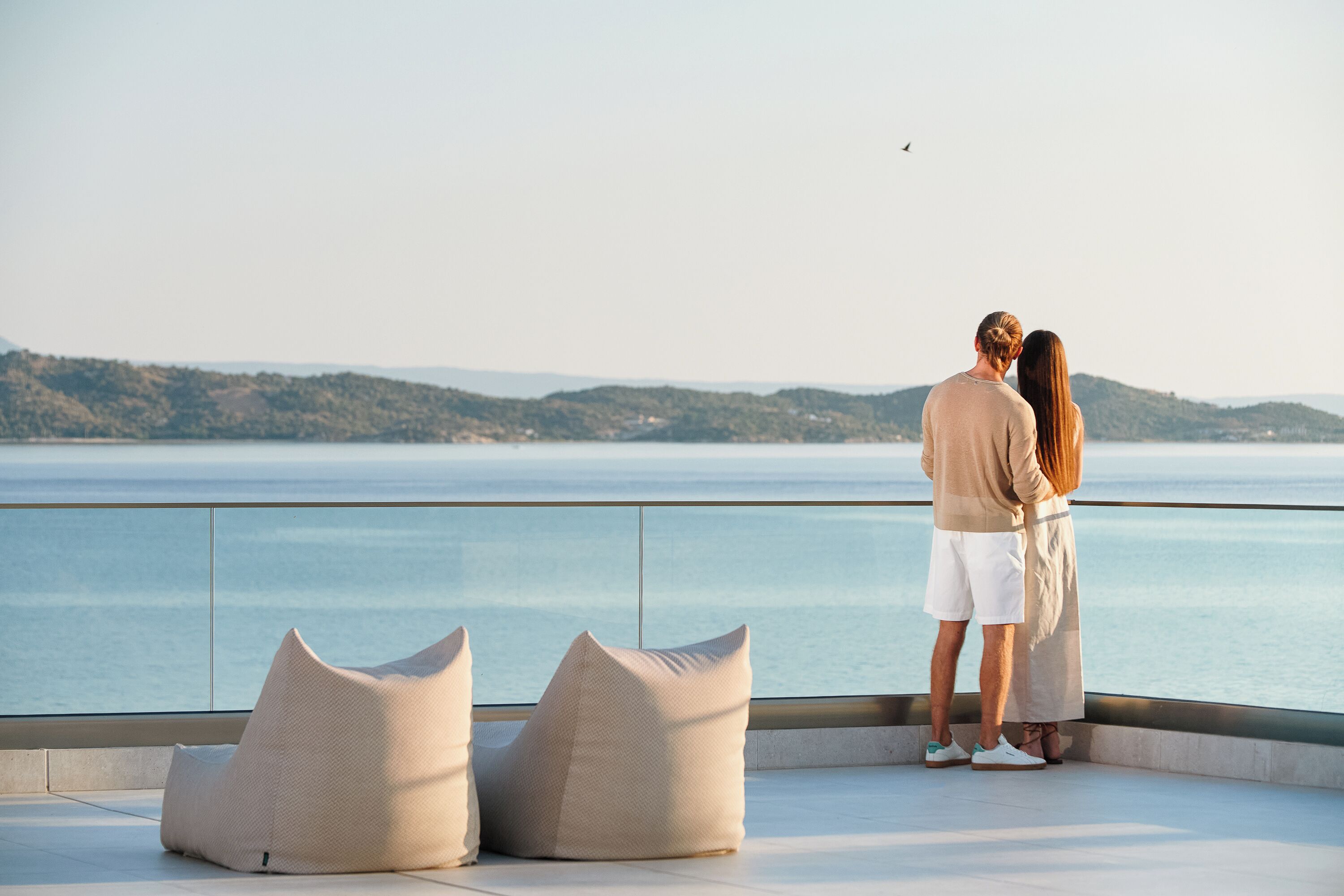 Una pareja de pie en un balcón con vista a un cuerpo de agua y colinas al fondo.