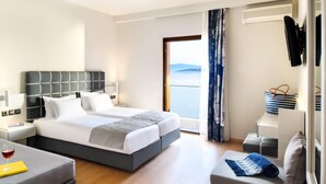 Quarto de hotel moderno com uma cama grande, uma mesinha de cabeceira com abajur, um pequeno espaço de estar e vista para o mar através de uma janela ampla.
