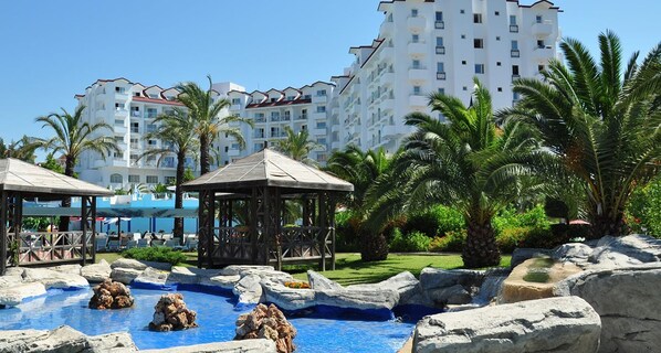 A resort with a swimming pool surrounded by rocks and palm trees, and a large hotel building in the background. - Serenis Hotel - All Inclusive (Manavgat)
