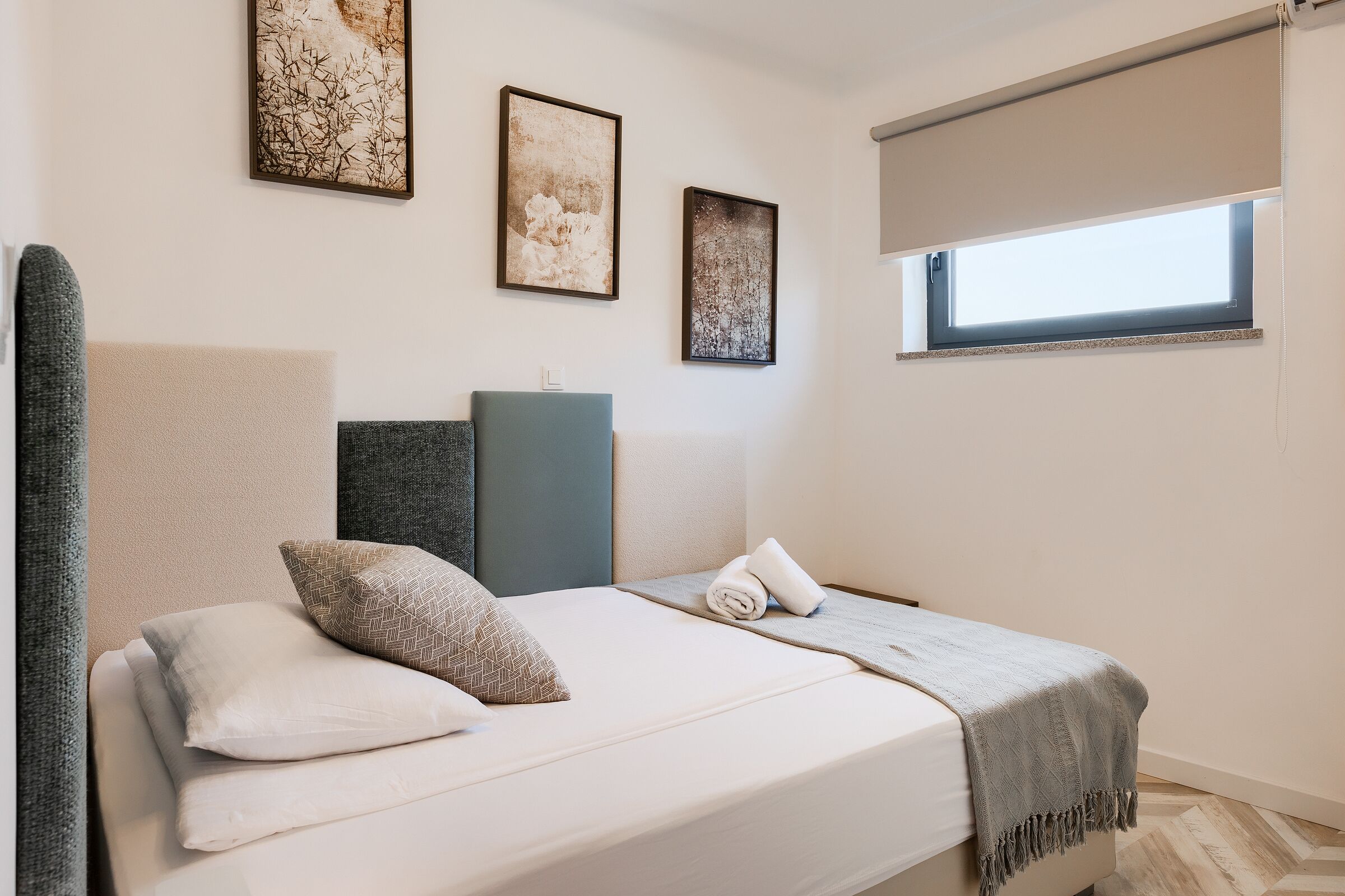 Una cama bien hecha con cabecera gris, una manta gris y una toalla enrollada. Encima de la cama hay dos cuadros enmarcados en la pared.