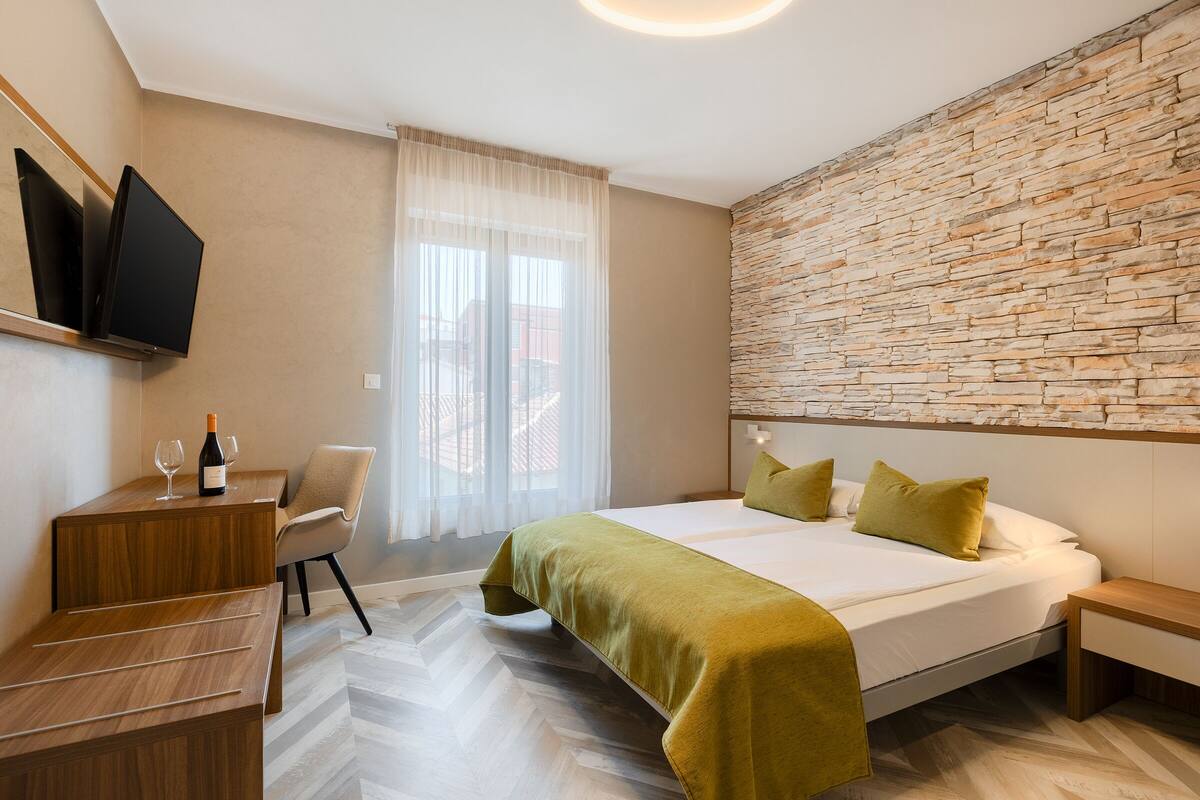 A hotel room with a bed, a desk with a bottle of wine and glasses, a chair, and a TV mounted on the wall.