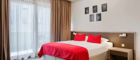 A modern hotel room with a large bed, a bedside table, and a headboard. There are curtains on the window, and artwork on the wall.