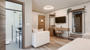 Une chambre d’hôtel moderne avec une douche en verre, un canapé blanc, un bureau, une chaise, un téléviseur fixé au mur et une porte en bois.