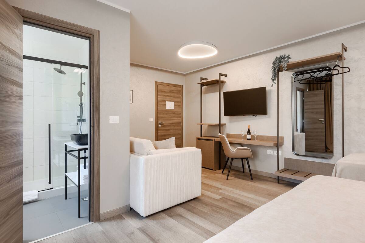 A modern hotel room with a glass shower, a white sofa, a desk, a chair, a TV mounted on the wall, and a wooden door.