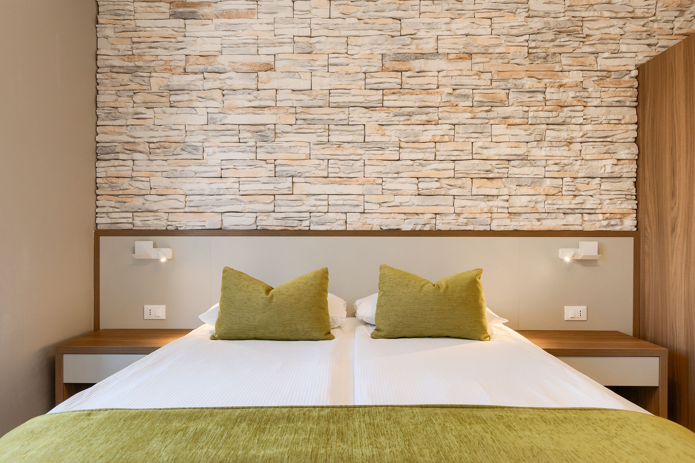 A bed with two pillows, a stone wall behind, and wooden nightstands with light switches.