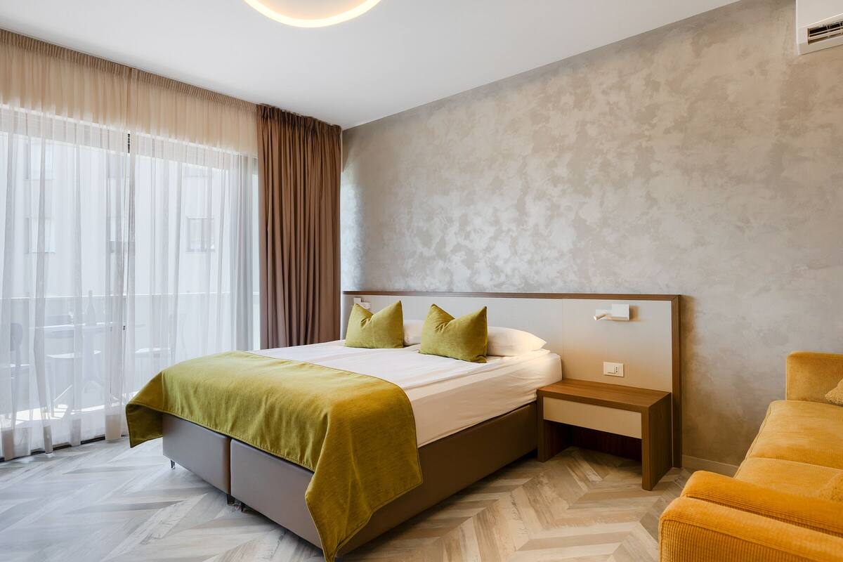 A modern hotel room with a large bed, a bedside table, a chair, and a view of the outdoors through sheer curtains.