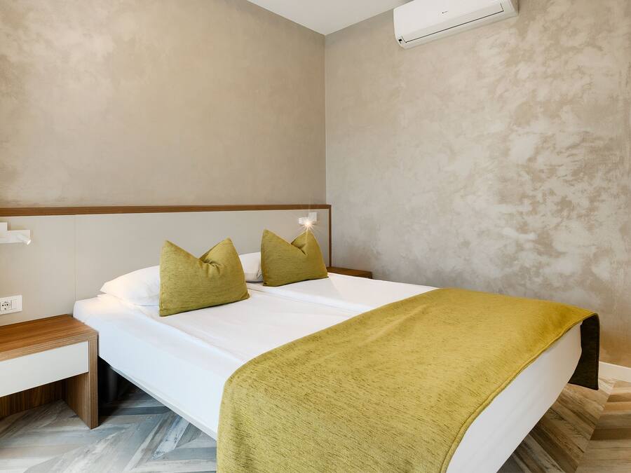Habitación de hotel con cama, mesita de noche, banco de madera y aire acondicionado de pared.
