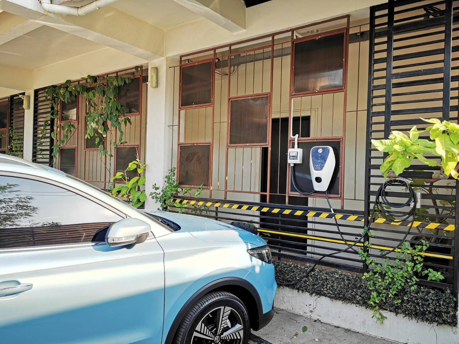 A blue car parked in front of a building with a black gate, a charging station, and a window with a plant.