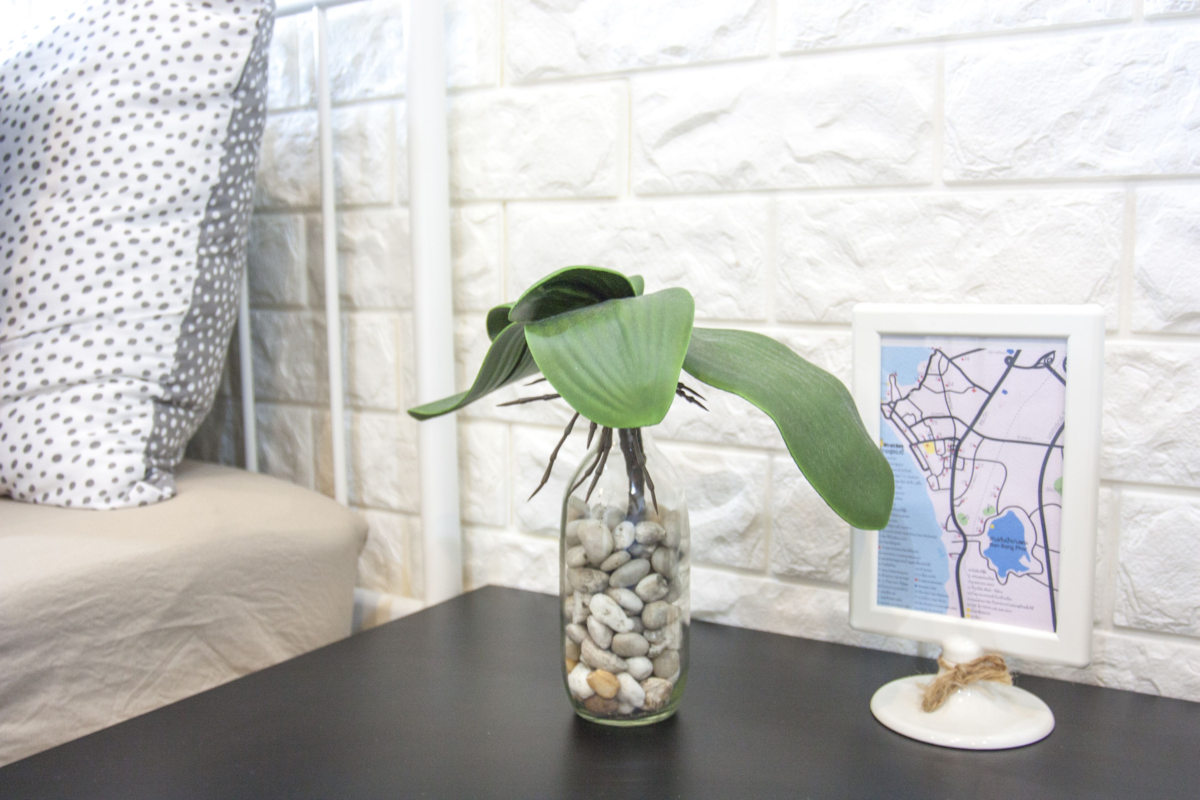 A glass vase with a plant on a dark surface, a white pillow with black dots, and a framed map on a white brick wall.