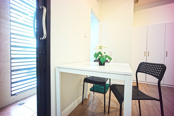 A room with a white table, a black chair, a green plant, and a window with blinds. - 26 Bed and Coffee (Chonburi)