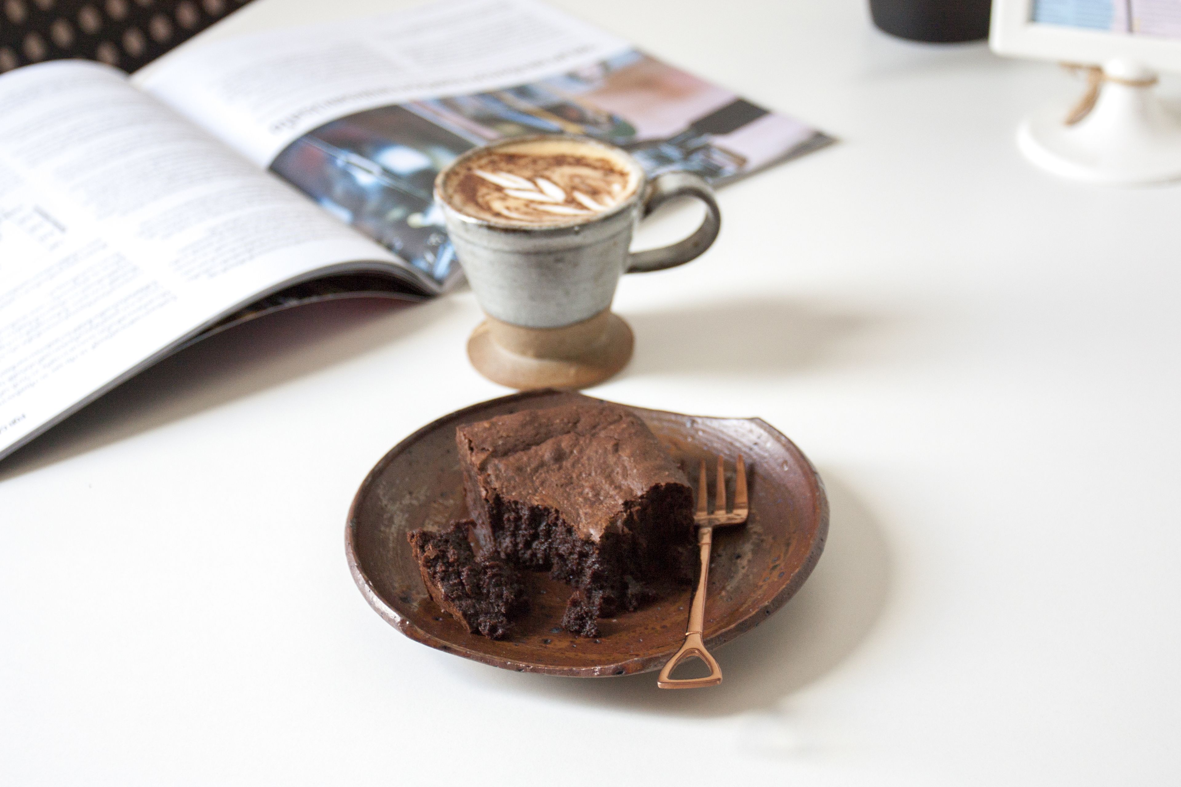 A slice of chocolate cake on a wooden plate with a fork beside it, a cup of coffee, and an open magazine on a white table.