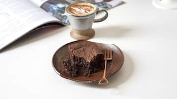 A slice of chocolate cake on a wooden plate with a fork beside it, a cup of coffee, and an open magazine on a white table.