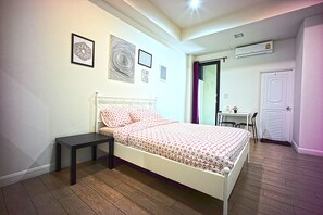 A bedroom with a white bed, a small bedside table, and a desk with a chair. There are framed pictures on the wall and an air conditioning unit above the desk. - 26 Bed and Coffee (Chonburi)