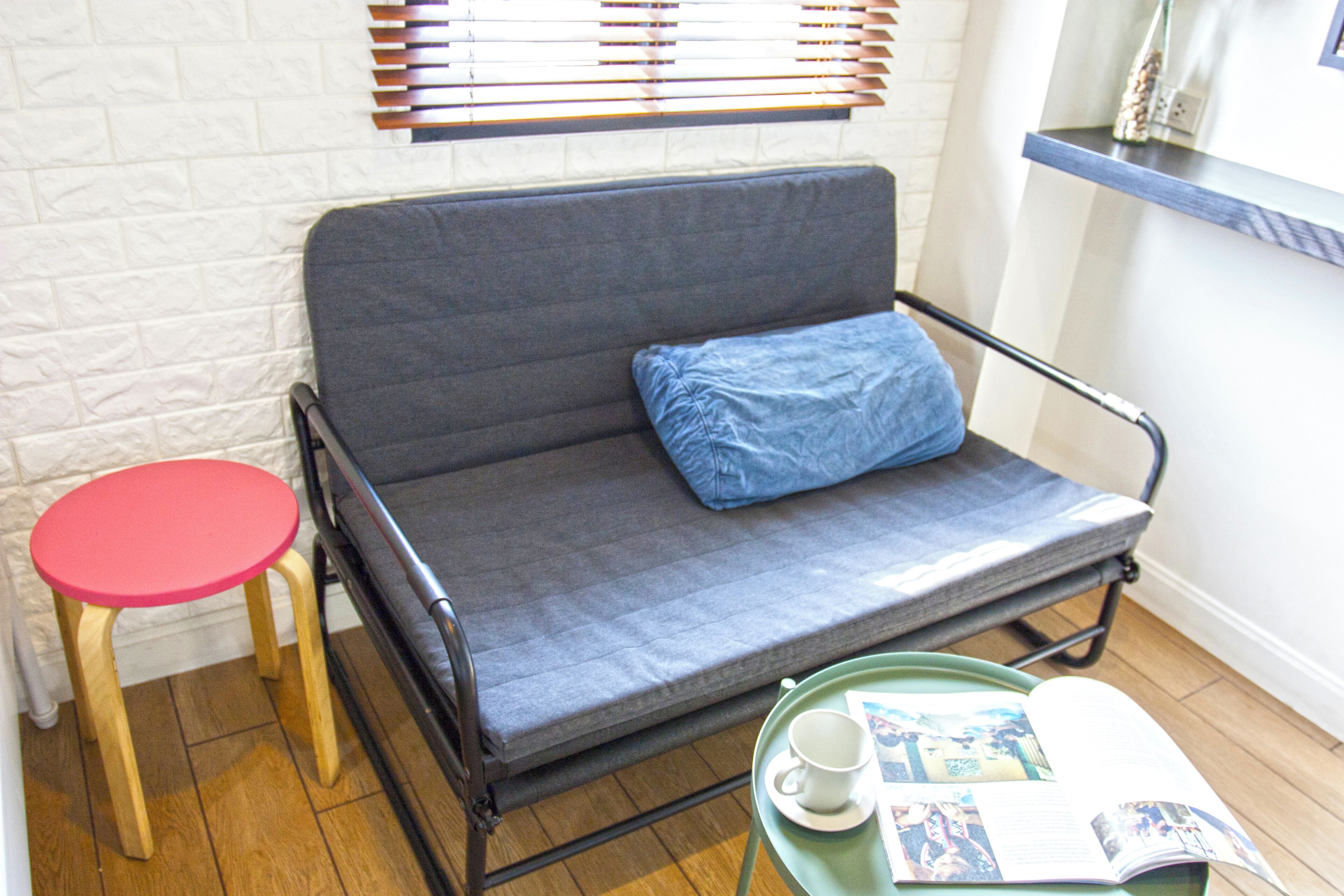 A small room with a grey sofa, a red stool, a green tray with a cup and magazine, and a white brick wall.