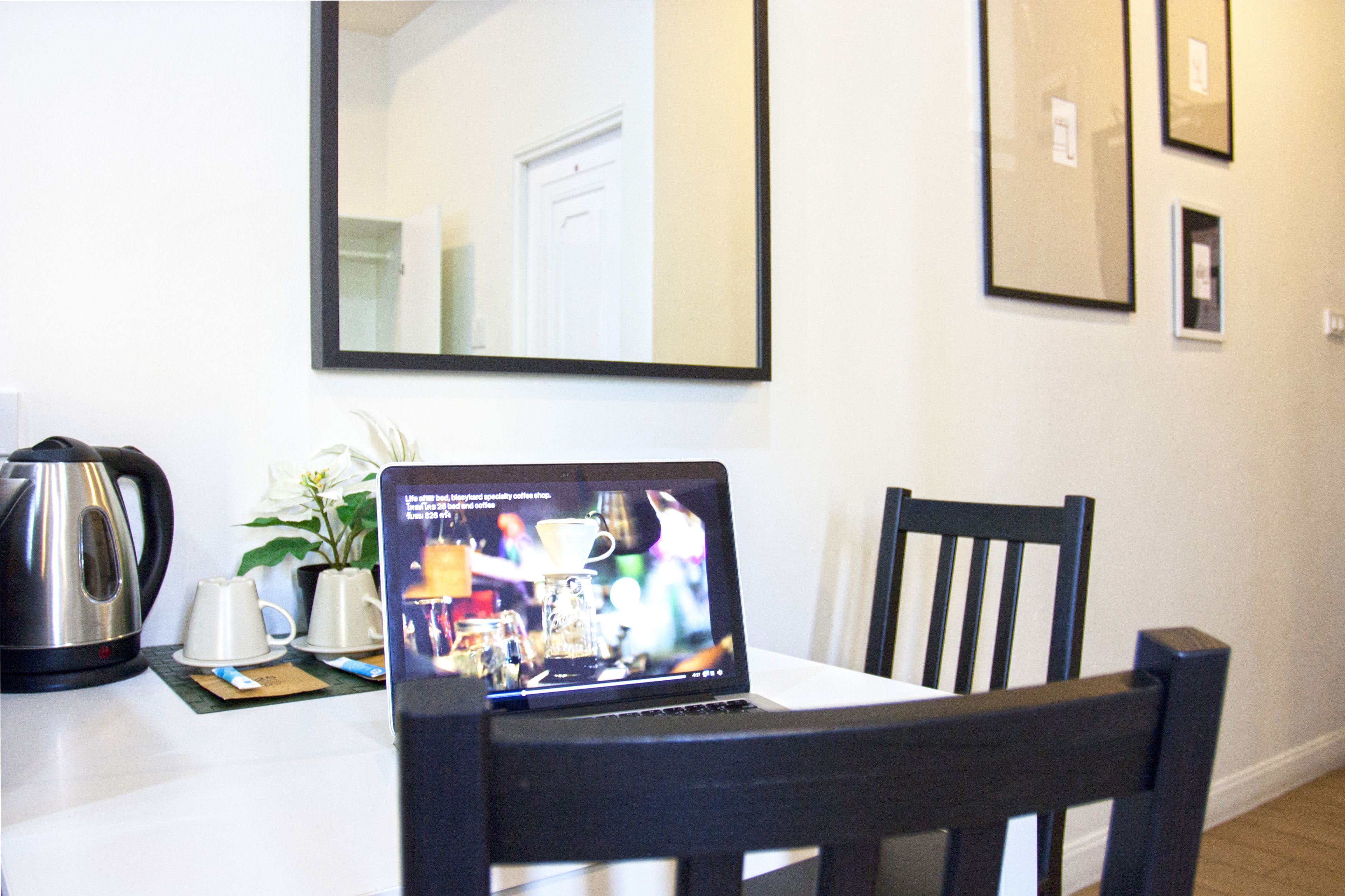 A laptop on a table with a coffee maker and cups, a plant, and framed pictures on the wall.