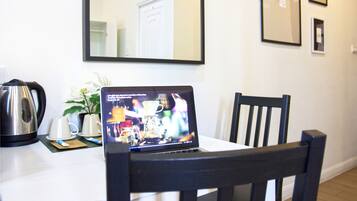 A laptop on a table with a coffee maker and cups, a plant, and framed pictures on the wall.