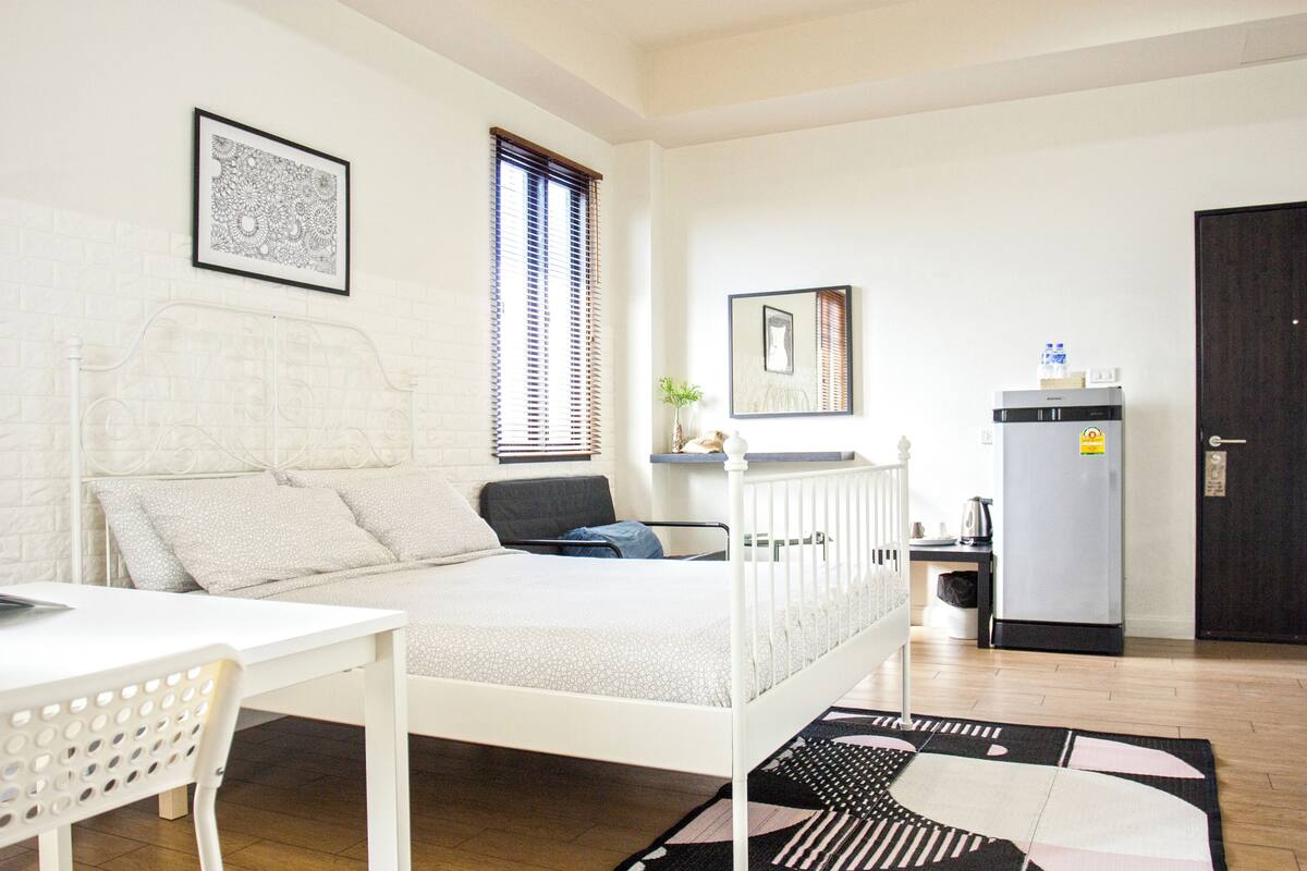 Un dormitorio con cama, escritorio, silla, refrigerador y puerta.