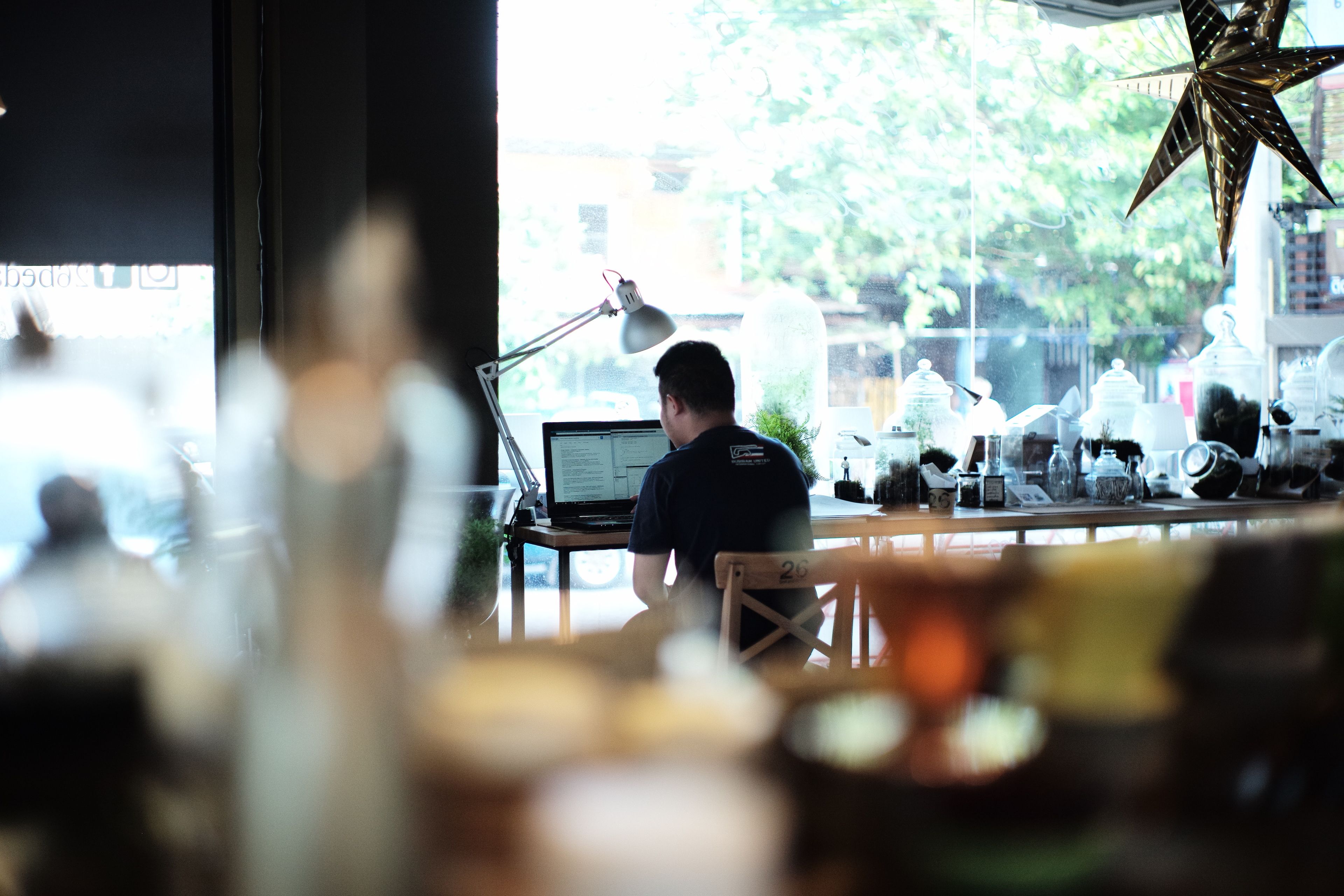 Eine Person arbeitet an einem Schreibtisch mit einem Laptop, einer Lampe und einer Stern-Dekoration in einem Zimmer mit einem Fenster.