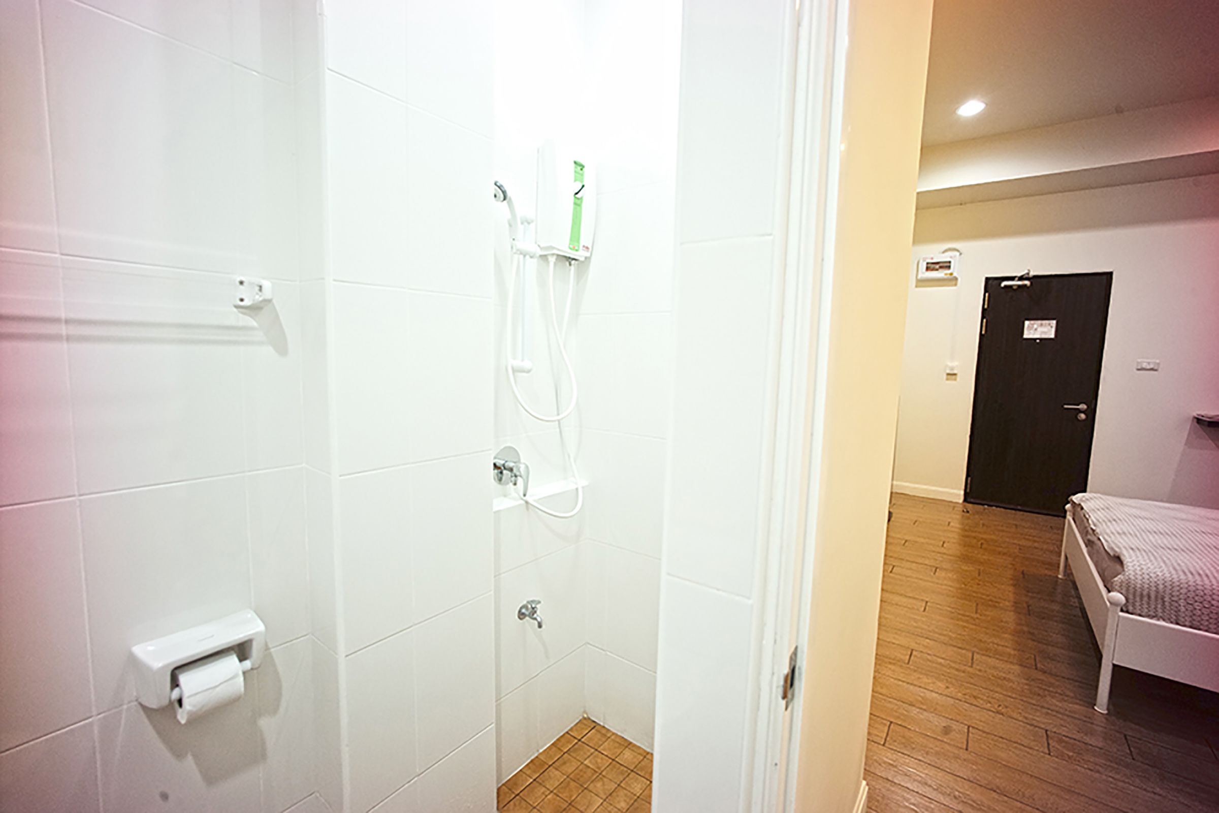 A small, clean bathroom with white tiles, a shower, a toilet, and a bed in the adjacent room.