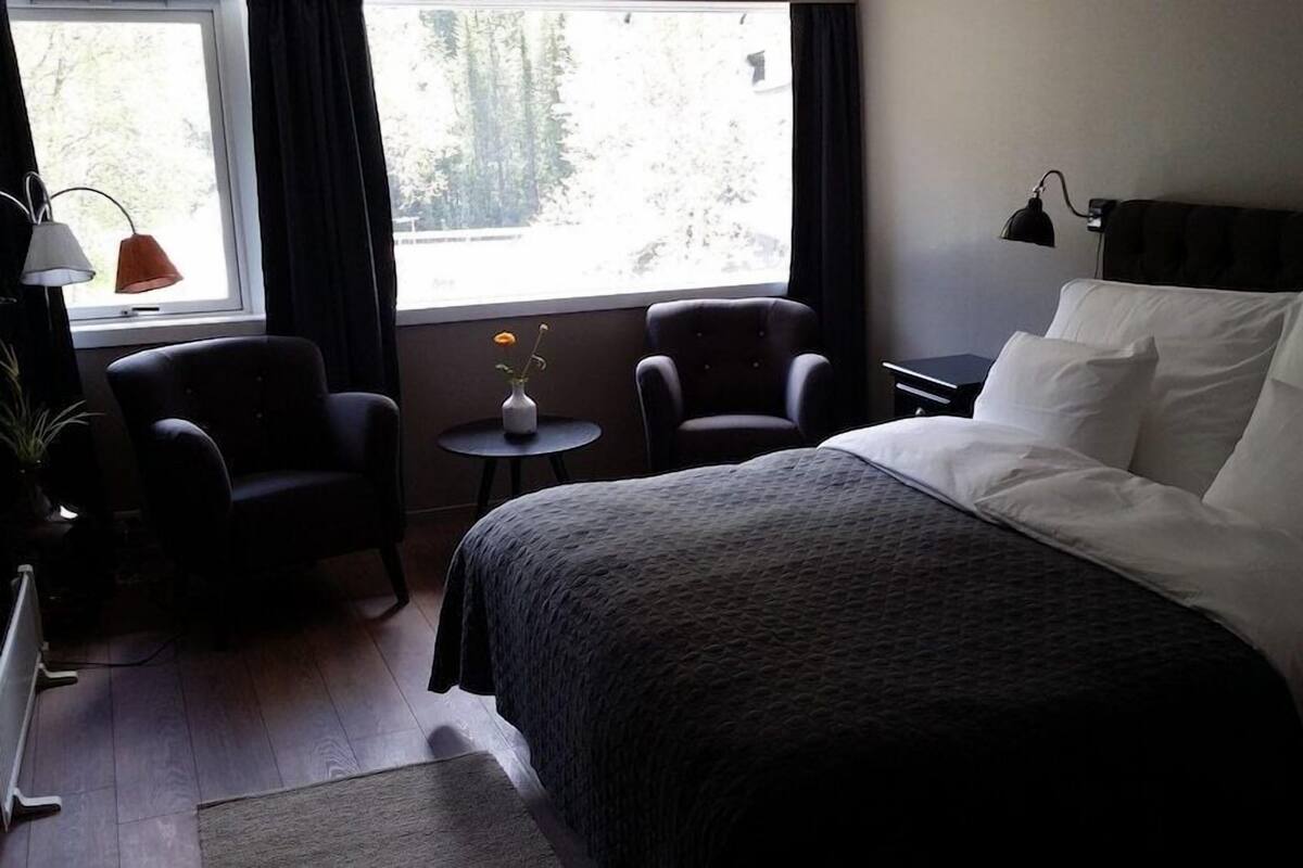 Een hotelkamer met een bed, twee fauteuils, een tafeltje met een vaas en een raam met gordijnen.